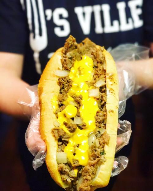 A person holds a large sandwich filled with shredded meat, onions, and melted cheese, wrapped in plastic.
