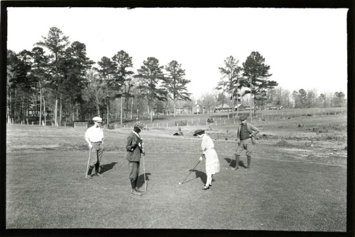 A black and white photo of four people playing golf on a grassy field, surrounded by trees and a distant house.