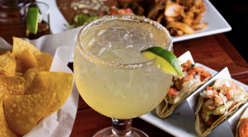 A refreshing margarita with a lime wedge, surrounded by chips, tacos, and a plate of food in the background.