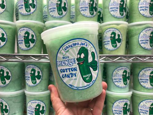 A hand holds a tub of green cotton candy labeled "Dilly Pickle Cotton Candy" with a cartoon pickle character.
