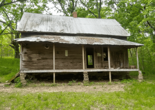 An old, weathered wooden house with a metal roof, surrounded by lush greenery and trees.