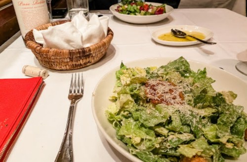 A table setting featuring a Caesar salad, a basket of bread, and a side salad with dressing in a restaurant.