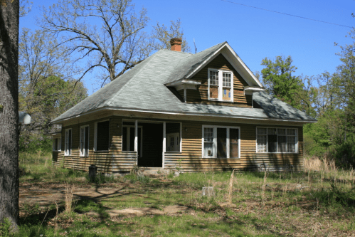 An abandoned two-story house with a green roof, surrounded by overgrown grass and trees. Windows are boarded up.