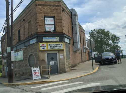 A brick building with a "We're OPEN" sign, featuring a Pepsi logo and a parked car nearby.