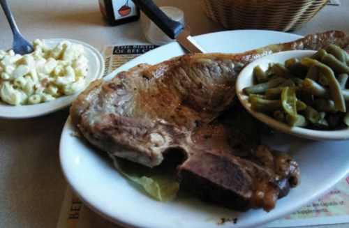 A large cooked steak on a plate, accompanied by green beans and macaroni salad in separate bowls.