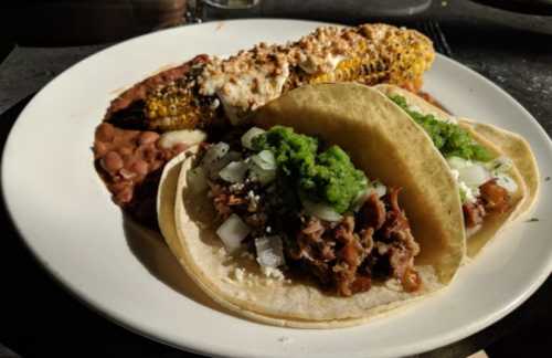 A plate featuring two tacos with meat and green sauce, alongside grilled corn and refried beans.