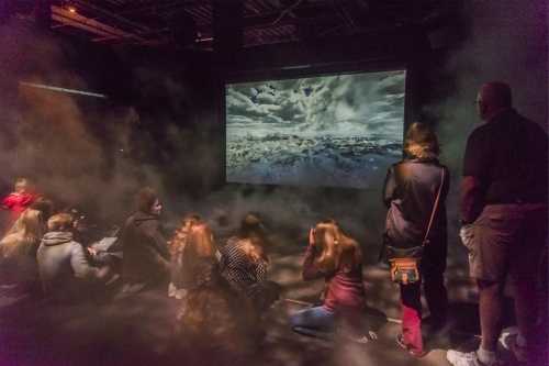 A group of people sits on the floor, watching a large screen displaying a dramatic landscape amidst a foggy atmosphere.