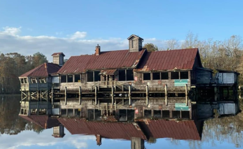 Abandoned wooden building with a red roof, reflecting in calm water, surrounded by trees under a clear blue sky.