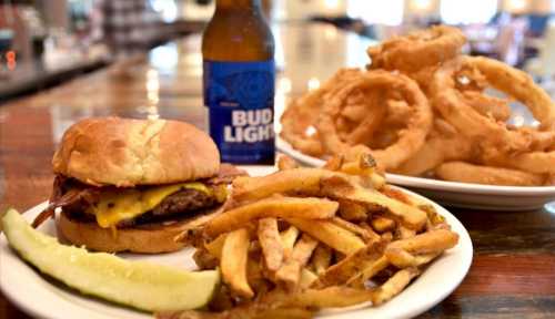 A plate with a cheeseburger, fries, onion rings, and a bottle of Bud Light on a wooden table.