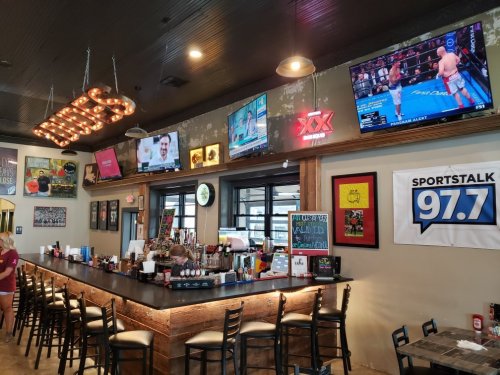 A lively bar interior with multiple TVs showing sports, neon signs, and a long counter with stools.
