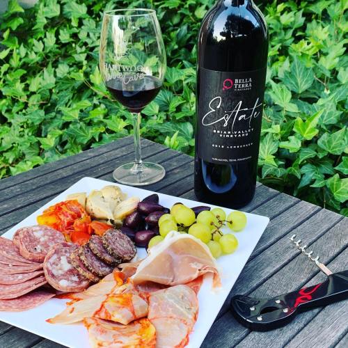 A bottle of red wine with a glass, accompanied by a charcuterie platter of meats, cheeses, and grapes on a wooden table.