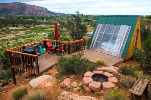A colorful cabin with a slanted roof on a deck, surrounded by desert landscape and a fire pit nearby.