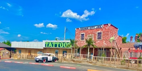 A colorful storefront featuring a tattoo shop and a jail, with a classic car parked outside under a blue sky.