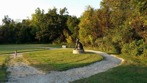 A child walks along a path towards a statue in a park, surrounded by trees and greenery.