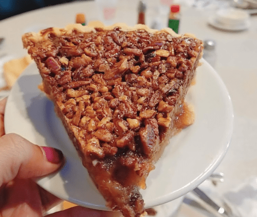 A hand holding a slice of pecan pie on a small plate, with a blurred background of a dining table.