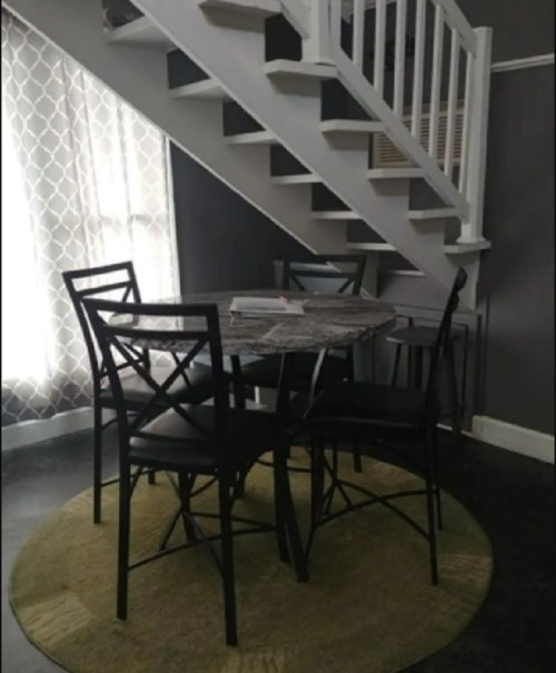 A small dining area with a round table and four black chairs, located under a staircase in a cozy room.