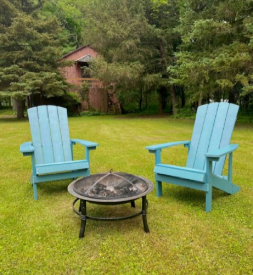 Two blue Adirondack chairs facing a fire pit on a grassy lawn, surrounded by trees and a rustic barn in the background.