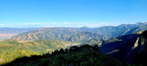 A panoramic view of lush green mountains under a clear blue sky, with valleys and ridges stretching into the distance.