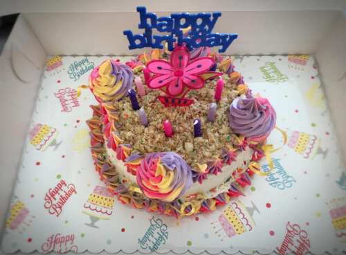 A colorful birthday cake with purple and pink frosting, candles, and a "Happy Birthday" topper in a decorated box.