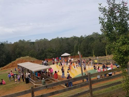 A large group of children and adults enjoying a colorful outdoor play area with a bouncy surface and trees in the background.