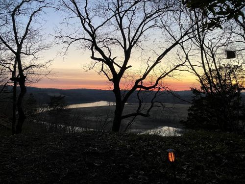 A serene sunset over a river, framed by bare trees and a peaceful landscape.
