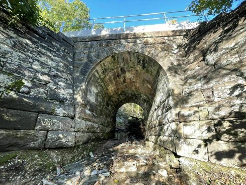 Stone arch bridge surrounded by greenery, with sunlight filtering through, viewed from below.