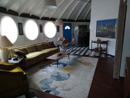 A modern living room with a round sofa, patterned rug, and large windows, featuring a dining area and stylish decor.