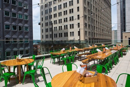 Rooftop dining area with wooden tables and green chairs, overlooking city buildings under a clear sky.