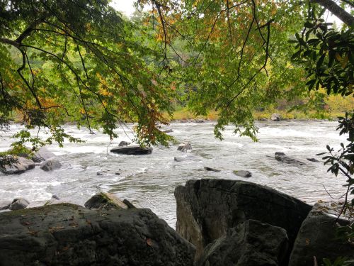 A serene river flows through a rocky landscape, framed by lush green trees and hints of autumn colors.