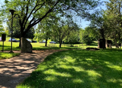 A sunny park scene with green grass, trees, and a stone path leading to picnic areas in the background.