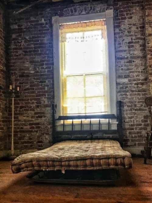 A simple, unmade bed with a plaid mattress sits in a rustic room with exposed brick walls and a bright window.