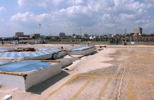 A view of a damaged rooftop with blue panels, overlooking a harbor and city skyline under a cloudy sky.