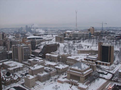 A snowy cityscape featuring buildings, a stadium, and distant skyline under a gray sky.