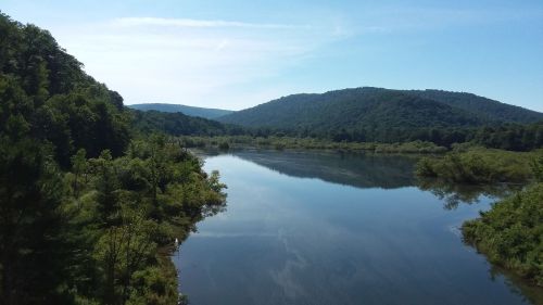 A serene river reflects the blue sky, surrounded by lush green hills and trees under bright sunlight.
