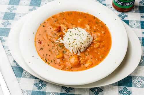 A bowl of shrimp gumbo with rice, garnished with herbs, on a checkered tablecloth. A bottle of garlic sauce is nearby.