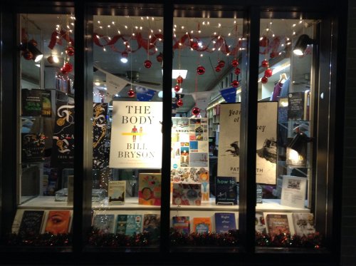 A bookstore window display featuring various books, holiday decorations, and a prominent sign for "The Body" by Bill Bryson.