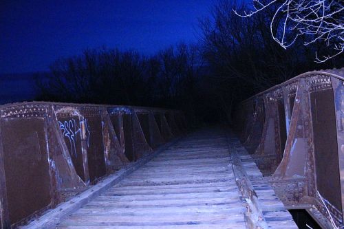 A dimly lit, abandoned bridge at night, surrounded by dark trees and a blueish sky. Graffiti is visible on the structure.