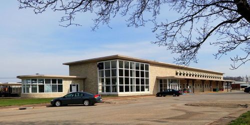 A modern building with large windows, surrounded by a parking lot and trees, under a clear blue sky.