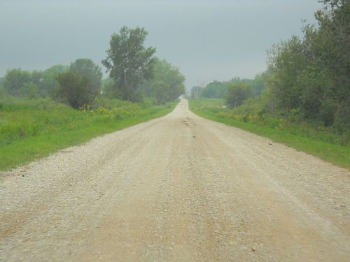 A gravel road stretches into the distance, flanked by greenery under a cloudy sky.