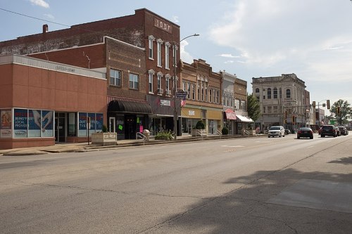 A quiet street lined with historic buildings and shops, with a few cars parked and passing by under a clear sky.