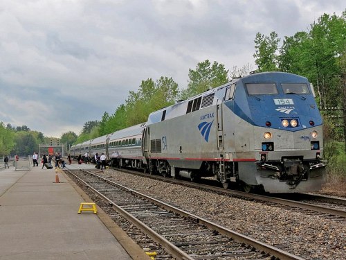 An Amtrak train at a station, with passengers boarding and trees in the background under a cloudy sky.