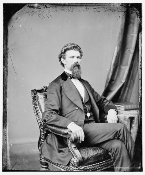 A seated man in a formal suit with a beard, posed elegantly in a vintage studio setting.