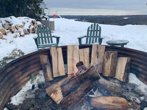 A cozy fire pit surrounded by snow, with logs stacked nearby and two chairs facing the flames against a winter landscape.