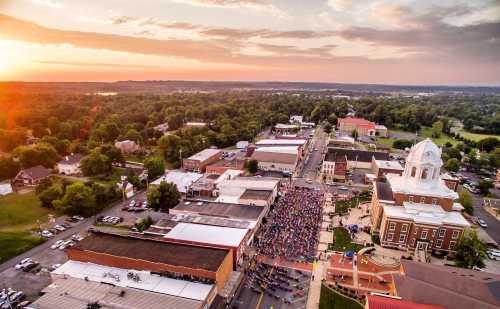 Aerial view of a bustling town square at sunset, with a crowd gathered and greenery surrounding the buildings.