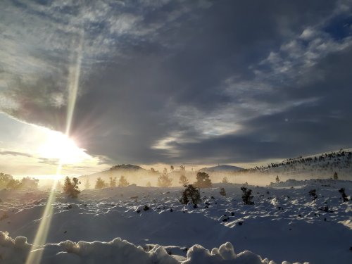 A snowy landscape with mist, illuminated by a bright sun breaking through clouds in the background.