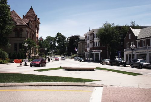 A quiet street scene featuring a roundabout, historic buildings, and parked cars under a clear blue sky.