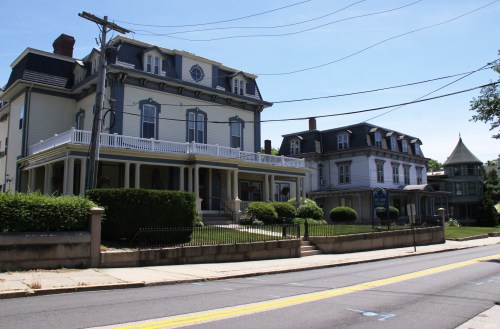 Two historic houses with porches and manicured lawns, set along a quiet street under a clear blue sky.