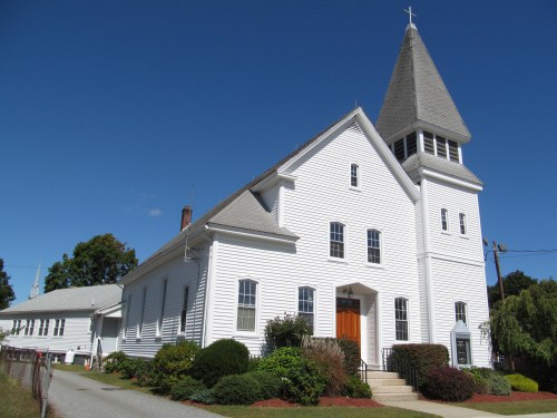 A white church with a tall steeple, surrounded by greenery and a clear blue sky.