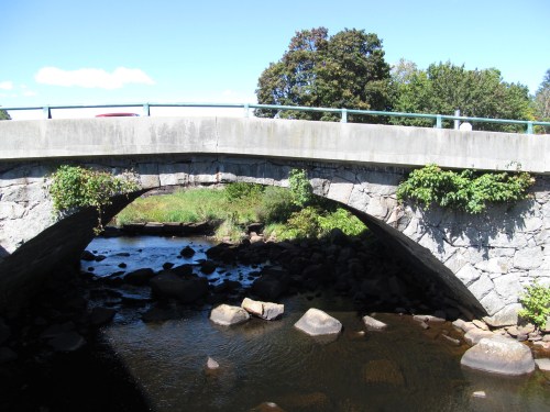 A stone arch bridge spans a calm river, surrounded by greenery and blue skies. Rocks are visible in the water below.