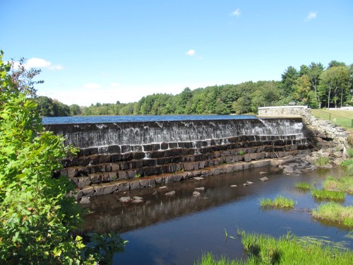 A serene view of a stone dam with cascading water, surrounded by greenery and a clear blue sky.
